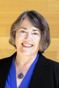 A headshot of a white woman with short brown gray hair wearing smiling and wearing a blue shirt with a black jacket and a blue pendant necklace.